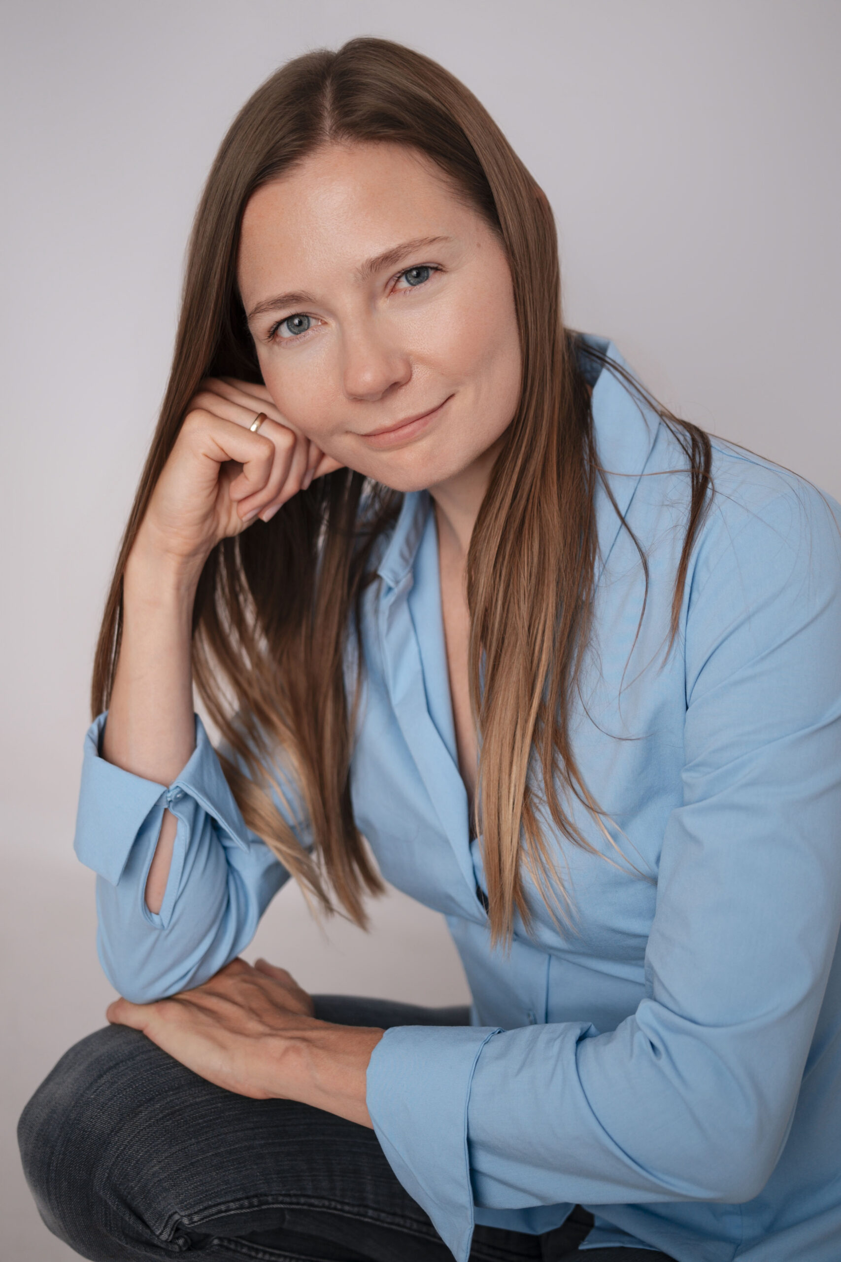 Portret siedzącej kobiety w jasnoniebieskiej koszuli, która opiera głowę na dłoni i patrzy w stronę aparatu